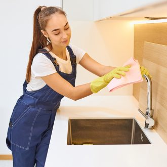 Young female worker of cleaning service wiping steel sink, wooden interior, modern